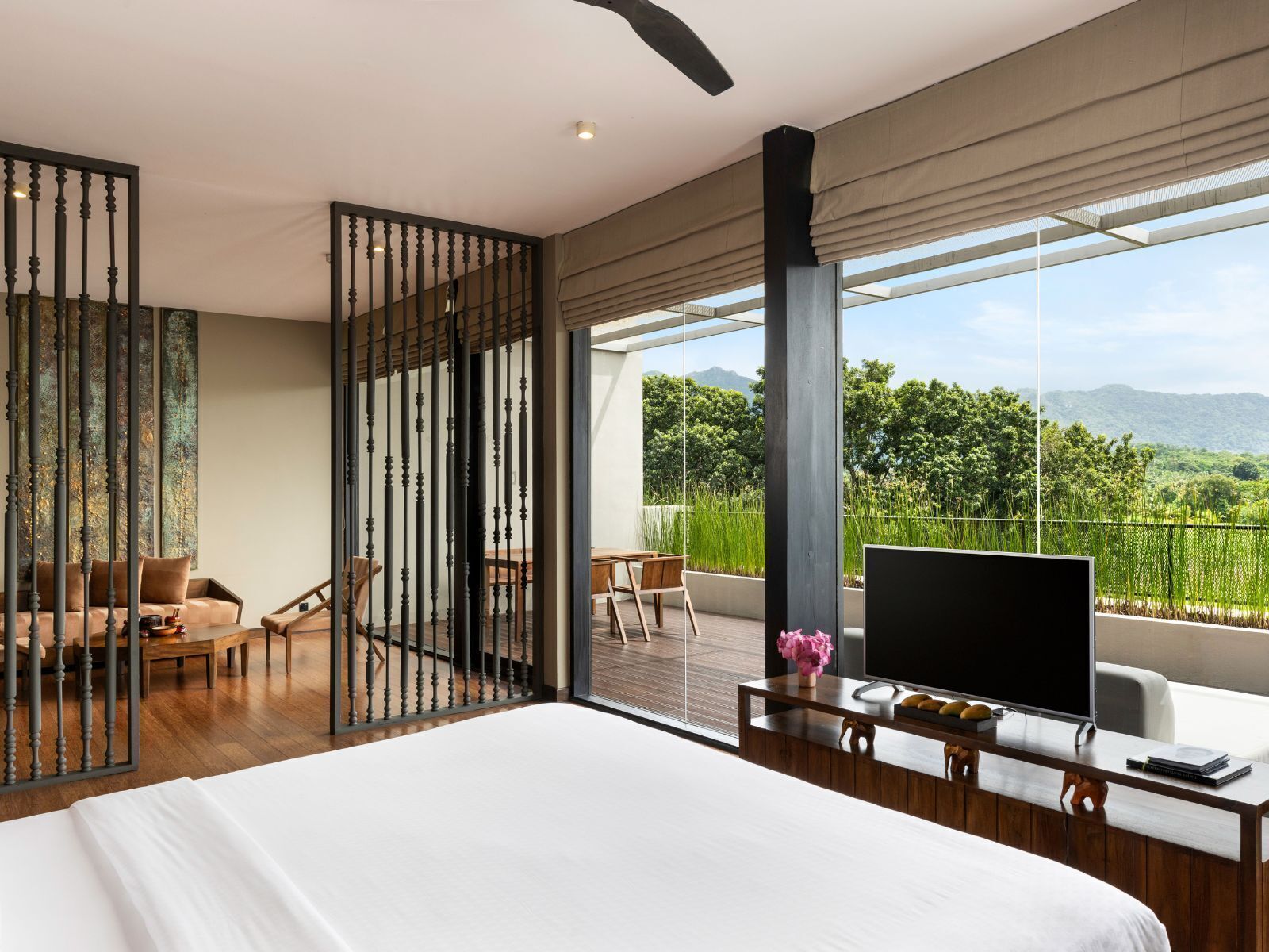 A modern hotel room with a large bed, a TV, a wooden desk, and a balcony with a view of greenery and mountains.