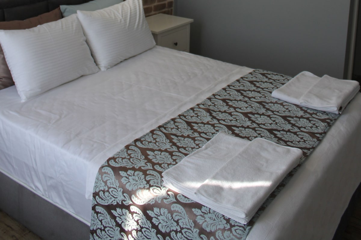 Una cama bien hecha con una colcha estampada, una lámpara colgante moderna y una pared de ladrillo.