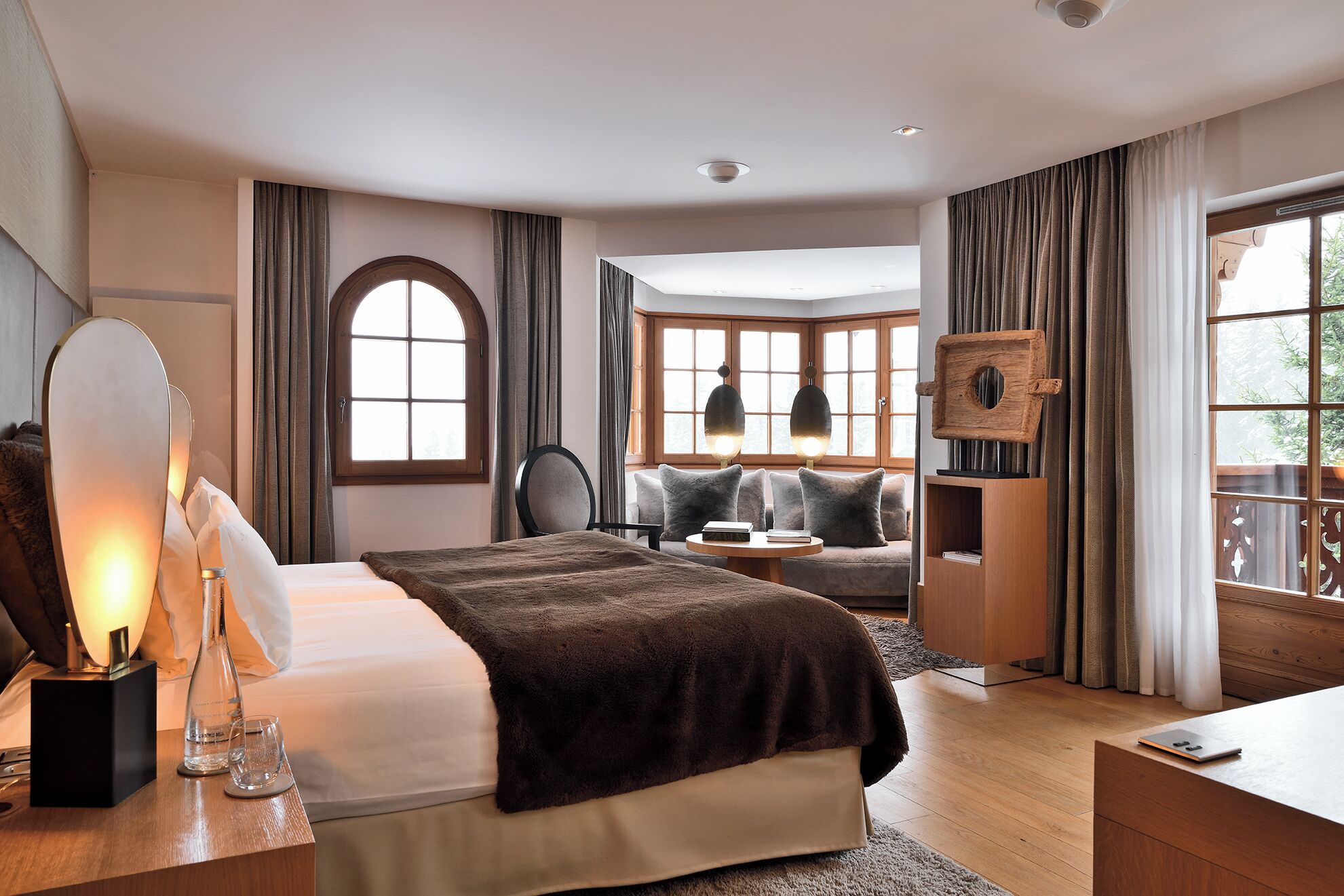 Un dormitorio moderno con una cama grande, una mesita de noche con una lámpara, una silla y un sofá. Se ven ventanas con cortinas, un cabecero de madera y una pequeña mesa con una lámpara.
