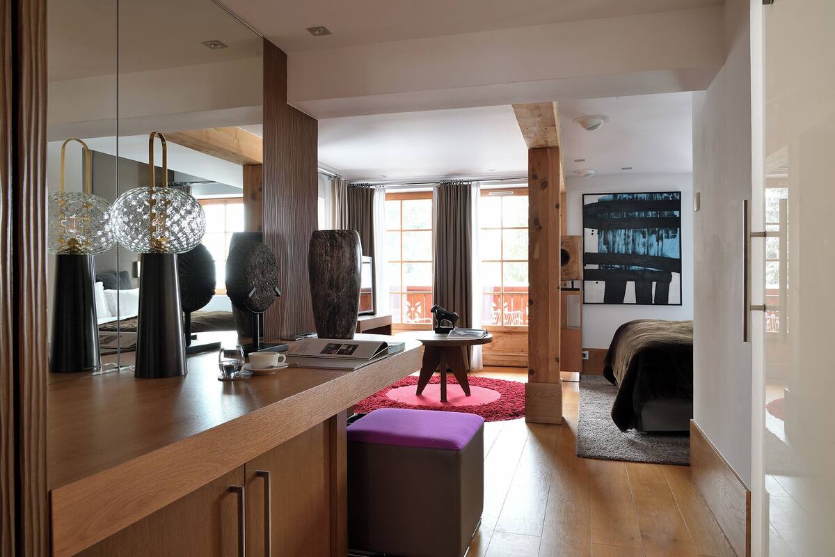 A modern hotel room with a wooden floor, a large mirror, a desk with a laptop, a bed with a red rug, and a large abstract painting on the wall.