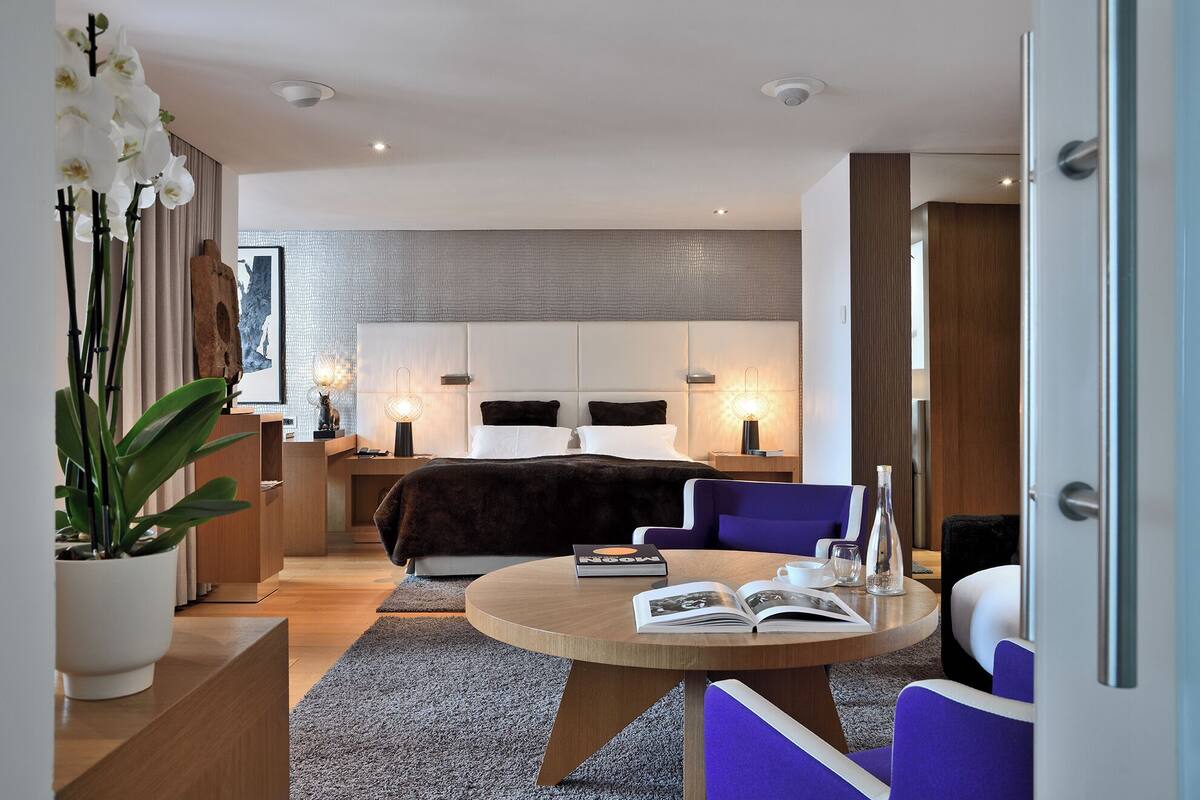 A modern hotel room with a bed, a round table with chairs, a potted plant, and a view of the city.