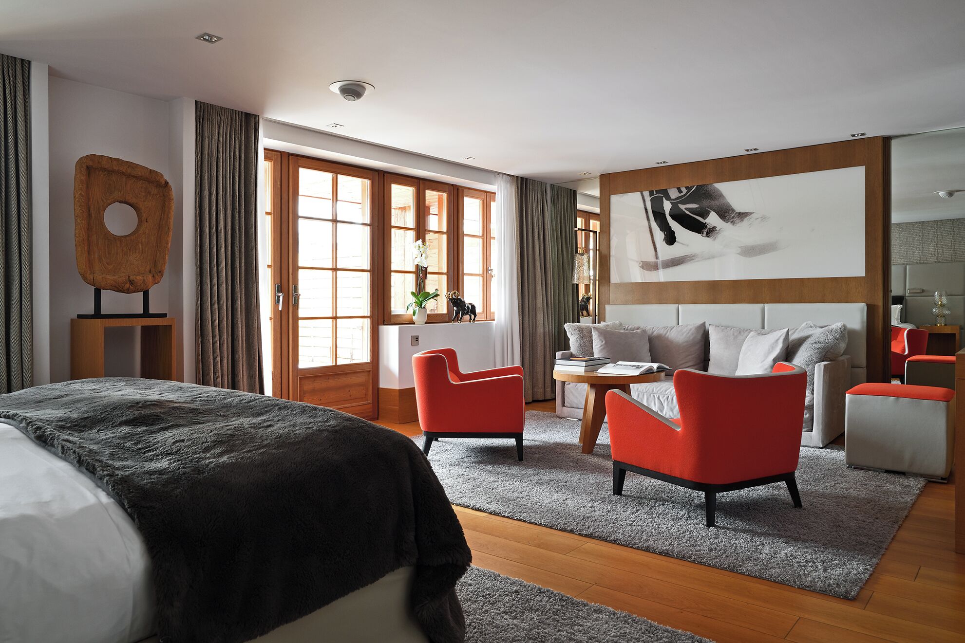 Une chambre d’hôtel moderne avec un grand lit, deux fauteuils rouges, un canapé gris, une tête de lit en bois et une grande fenêtre avec des rideaux.