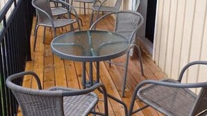 A wooden deck with a glass table and wicker chairs, overlooking a building with large windows and a door.