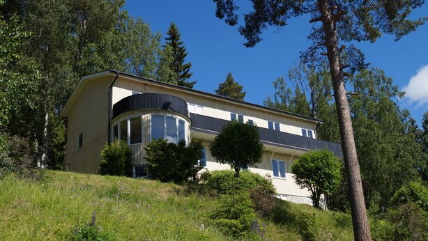 A two-story house with a curved balcony, surrounded by trees and a grassy hill.
