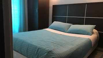 Un lit bien fait, avec une couvre-lit bleue et des oreillers, une tête de lit sombre et un luminaire fixé au mur.