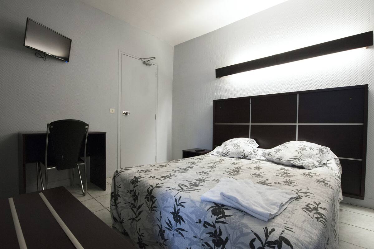 A bedroom with a bed, a desk, a chair, a TV, and a bedside table.