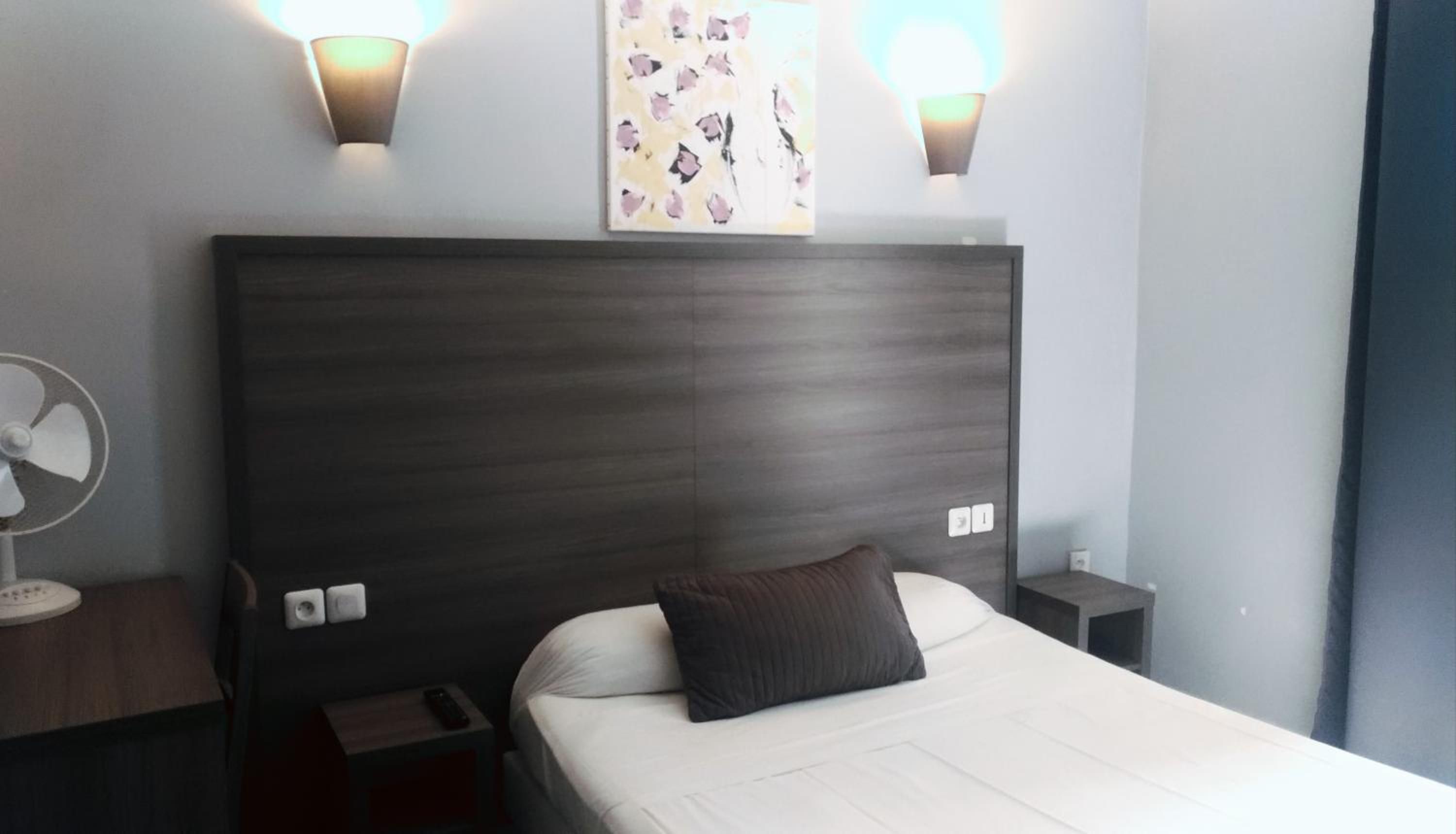 A hotel room with a large bed, a bedside table with a lamp, a wall-mounted light fixture, and a small nightstand.