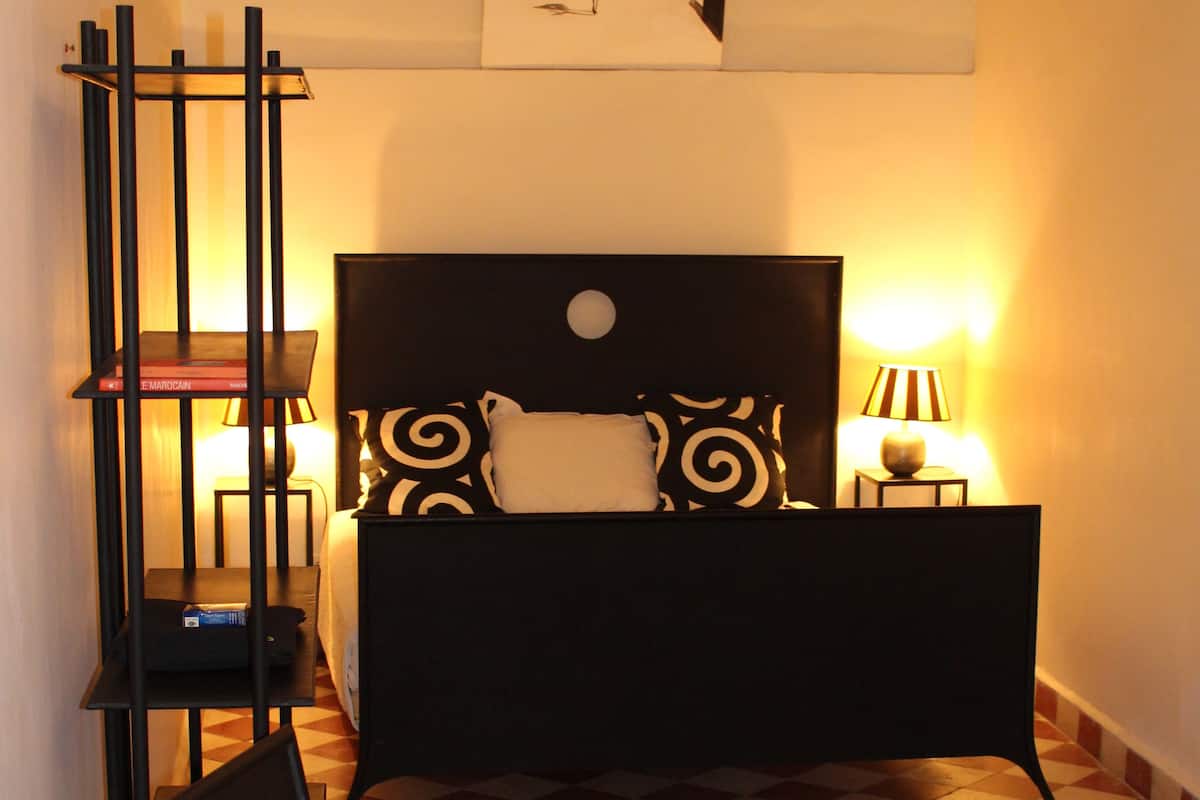 Un dormitorio con cama, una silla, una mesita de noche, una lámpara y una obra de arte en la pared.
