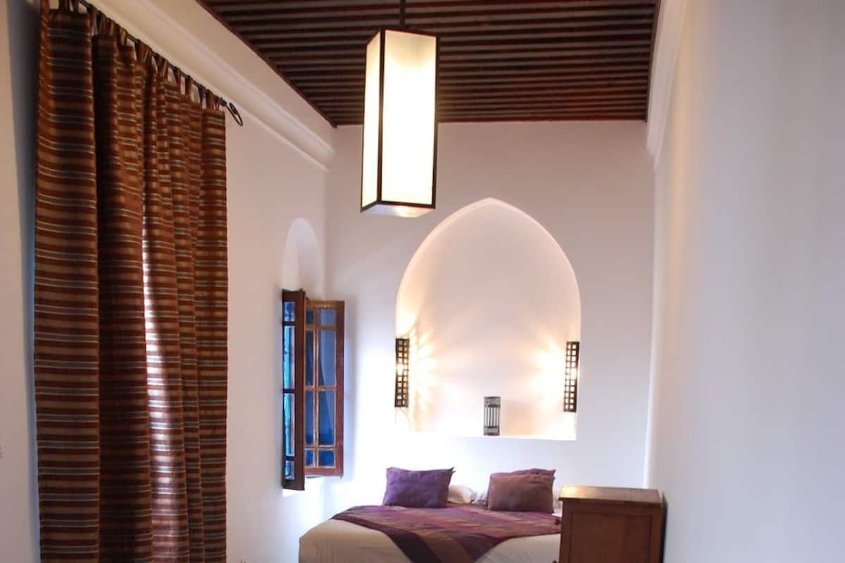 Un dormitorio con una cama, cabecera de madera, alfombra estampada y una ventana con cortinas.