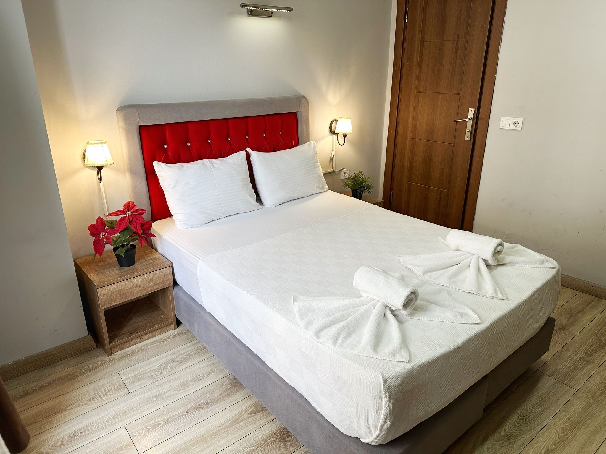 A neatly made bed with white linens and towels, a red upholstered headboard, bedside lamps, and a small potted plant on the nightstand.
