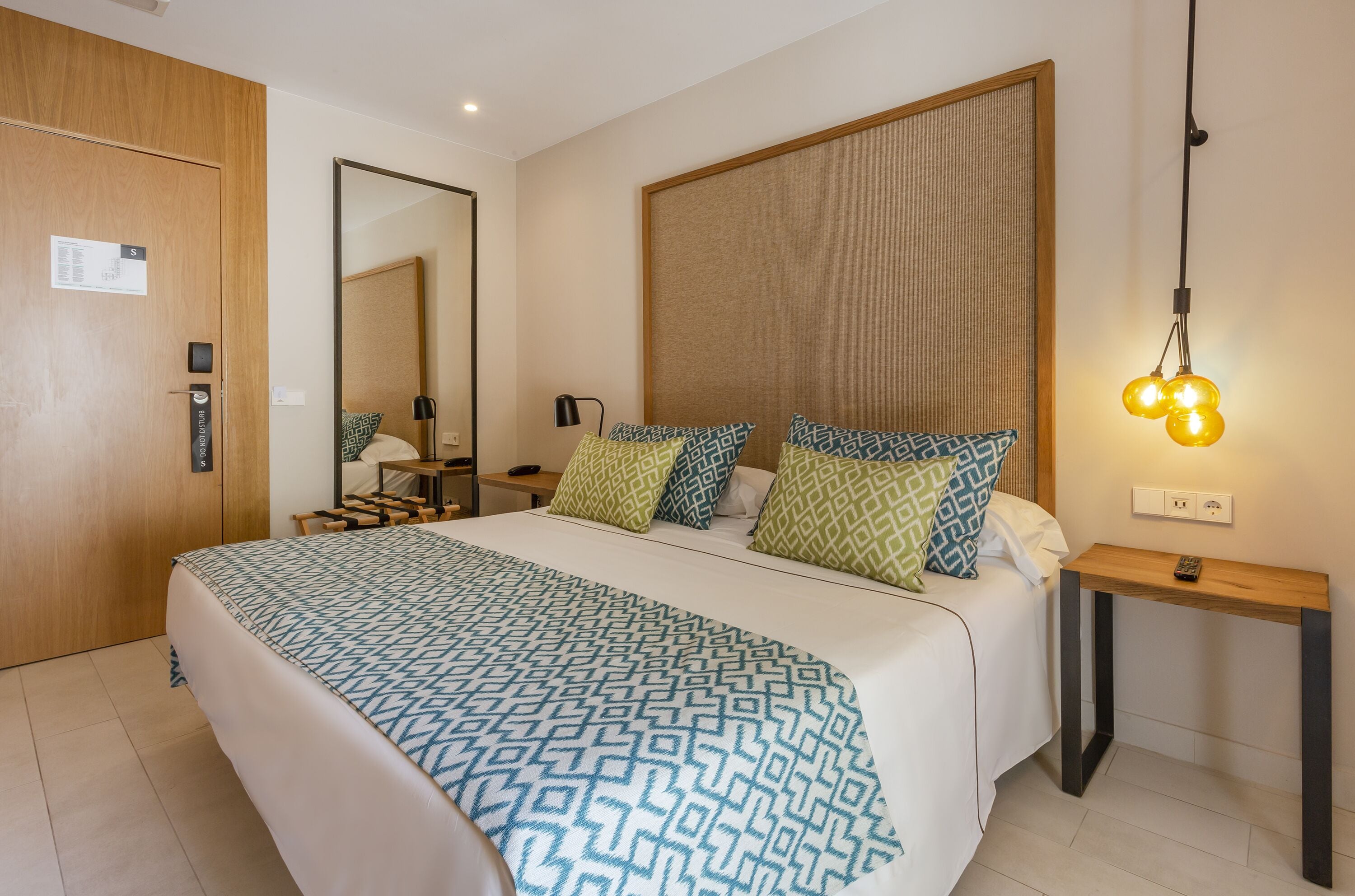 A hotel room with a large bed, a wooden door, a bedside table with a lamp, and a mirror on the wall.