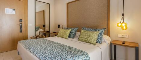 A hotel room with a large bed, a wooden door, a bedside table with a lamp, and a mirror on the wall.