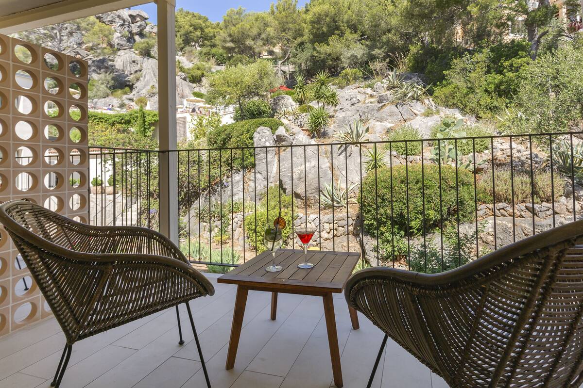 A balcony with wicker chairs and a table, overlooking a rocky garden with trees and a view of a building in the background.