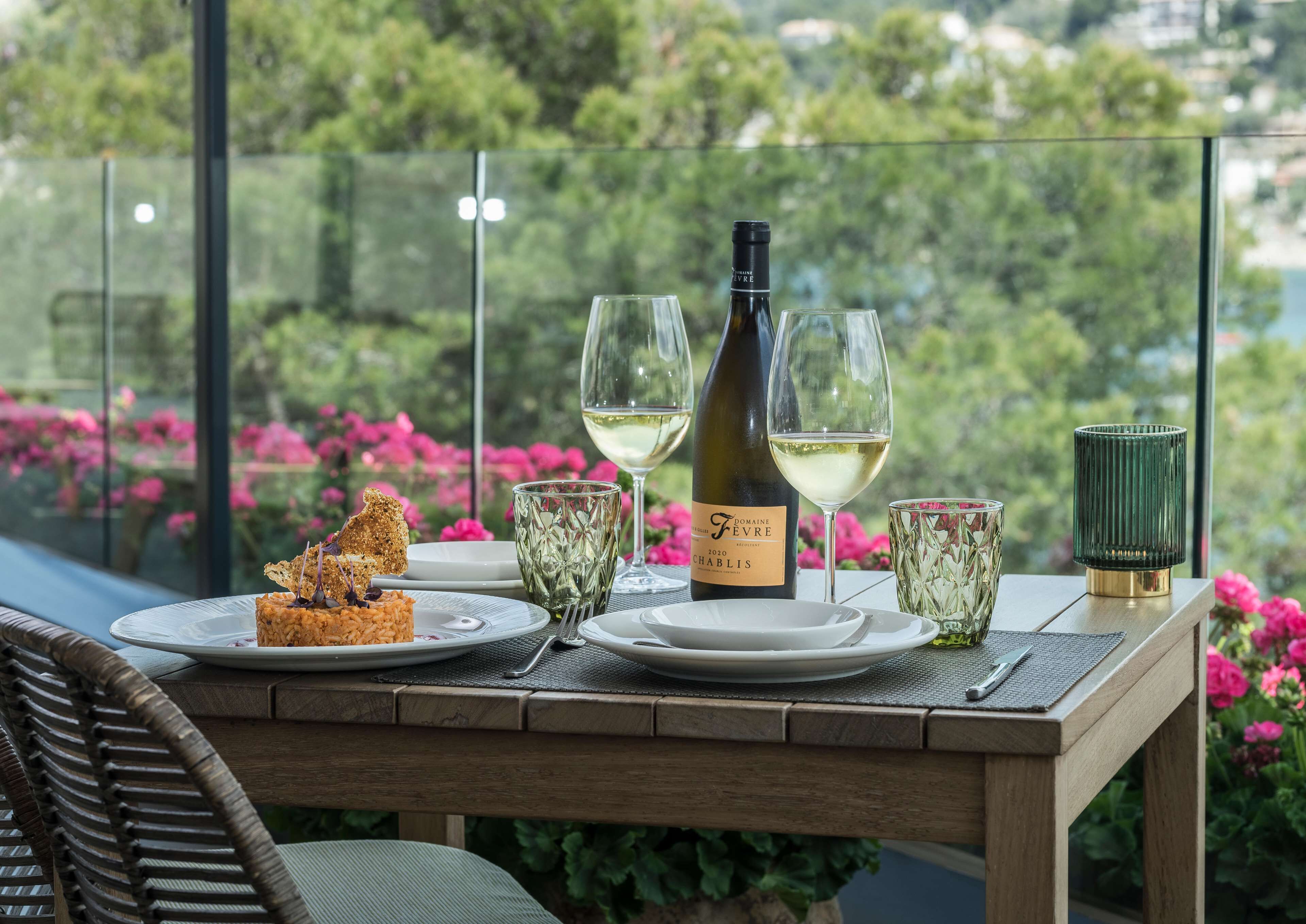 A table set with a bottle of wine, glasses, and a dessert on a balcony with a view of greenery and flowers.