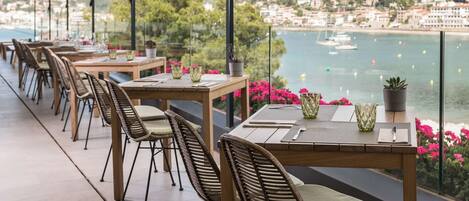 A terrace with wooden tables and wicker chairs, overlooking a coastal town and the sea.