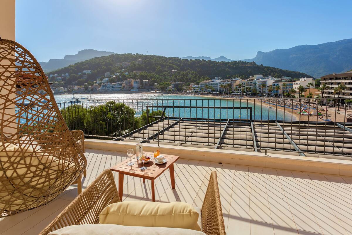 A balcony with wicker furniture, a table with a drink and snacks, and a view of a beach and mountains.