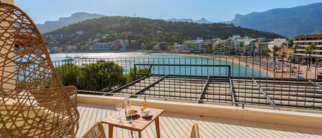 Un balcón con mobiliario de mimbre, una mesa con bebidas y aperitivos, y vistas a la playa y las montañas.