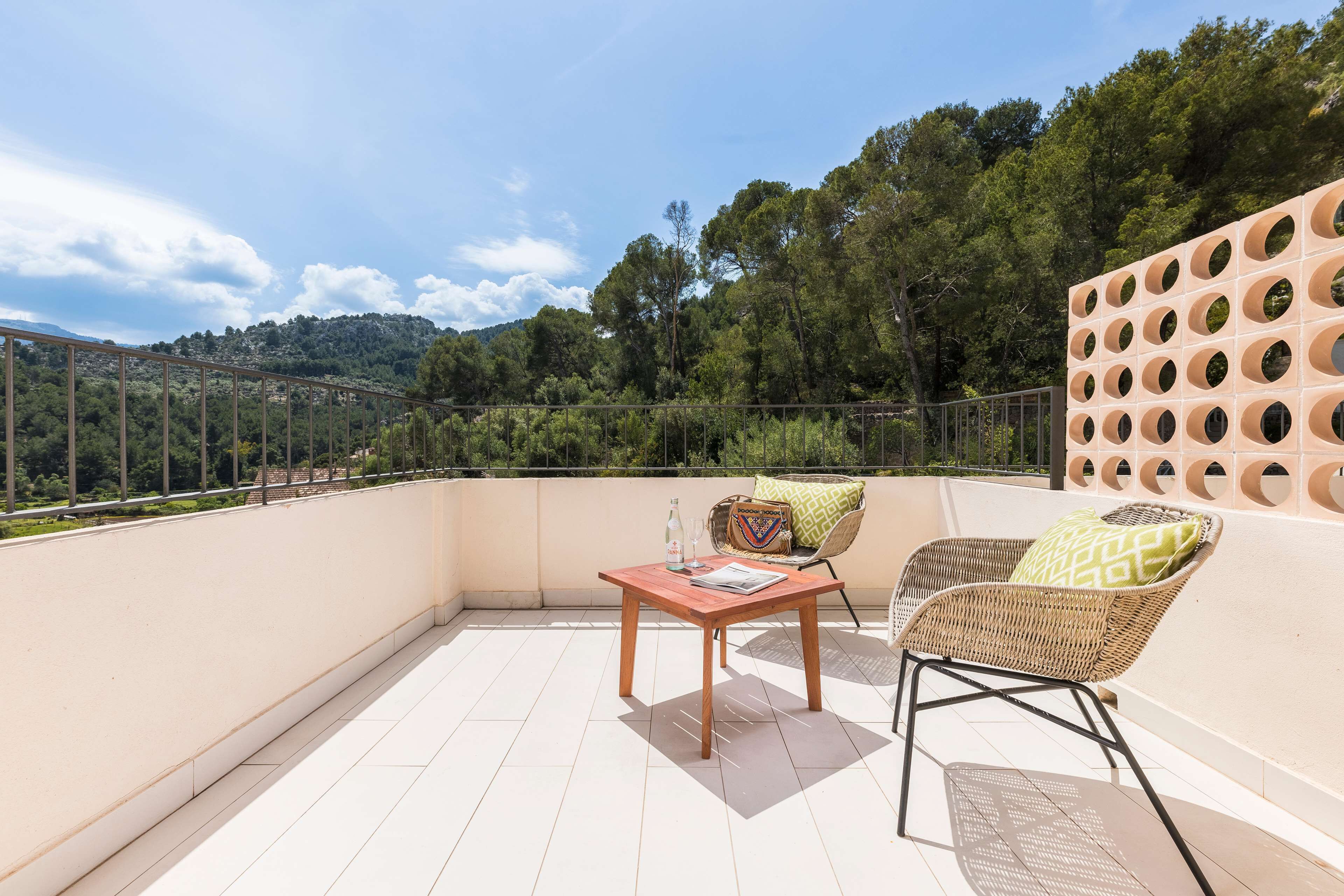 Een balkon met een tafel en twee rieten stoelen, waarvan er een een bekussen met patroon heeft.