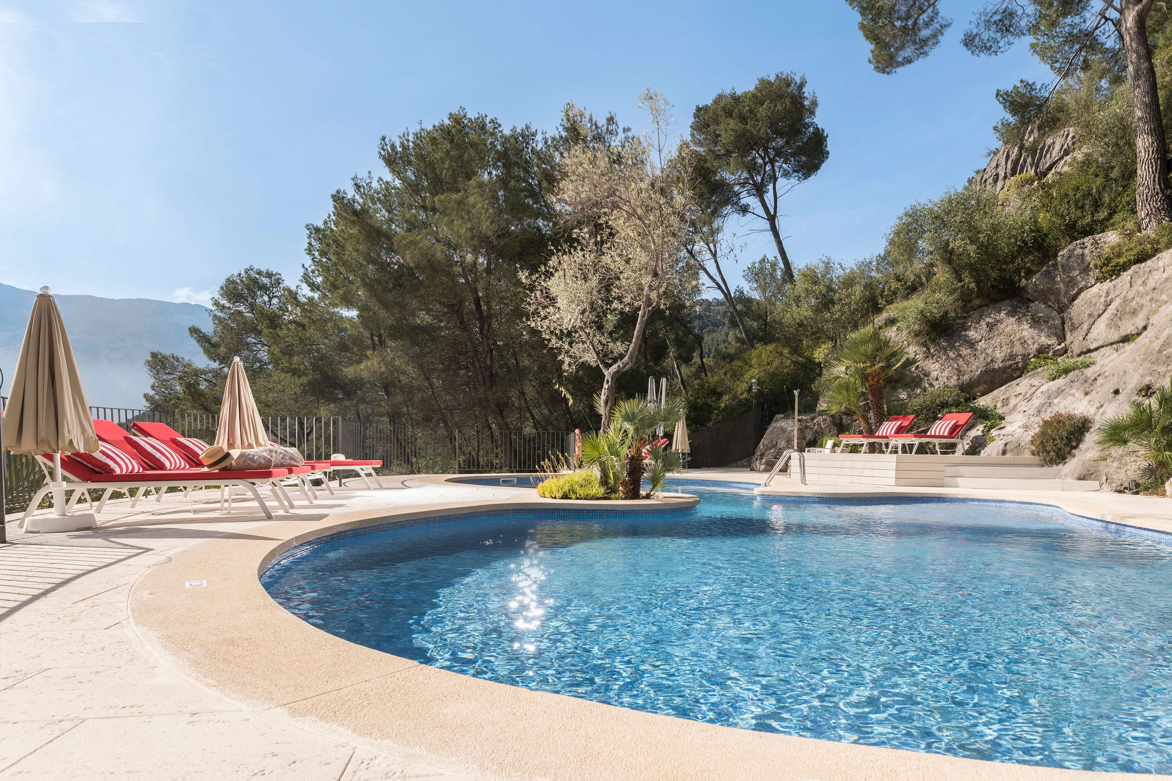 Una piscina con sillas de descanso rojas y blancas, rodeada de árboles y una ladera rocosa.