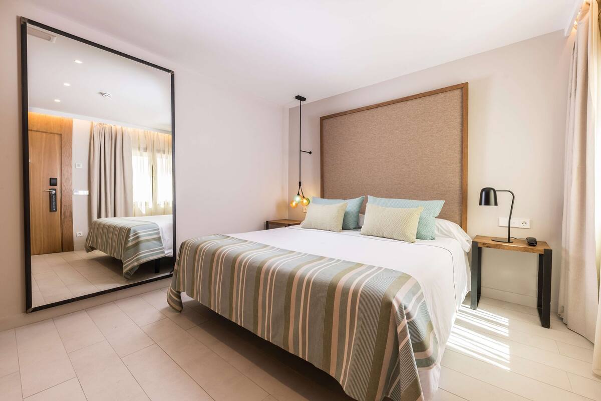 A hotel room with a large bed, a bedside table with a lamp, a mirror, and a wooden headboard.