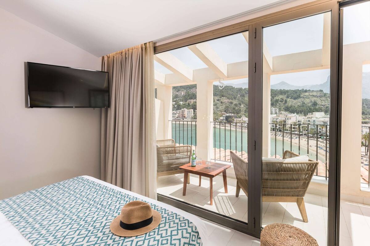 A hotel room with a bed, a TV mounted on the wall, a balcony with a view of the sea and buildings, and wicker furniture.