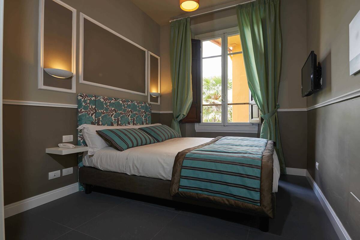 A bedroom with a bed, a bedside table, a wall-mounted TV, and a window with curtains.