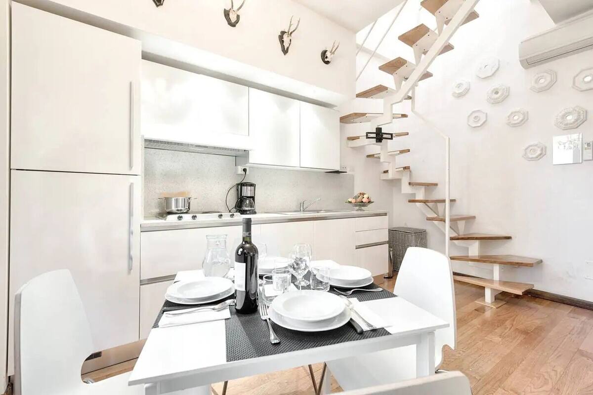 A modern kitchen with white cabinetry, a dining area with a table set for two, and a staircase with wooden steps.