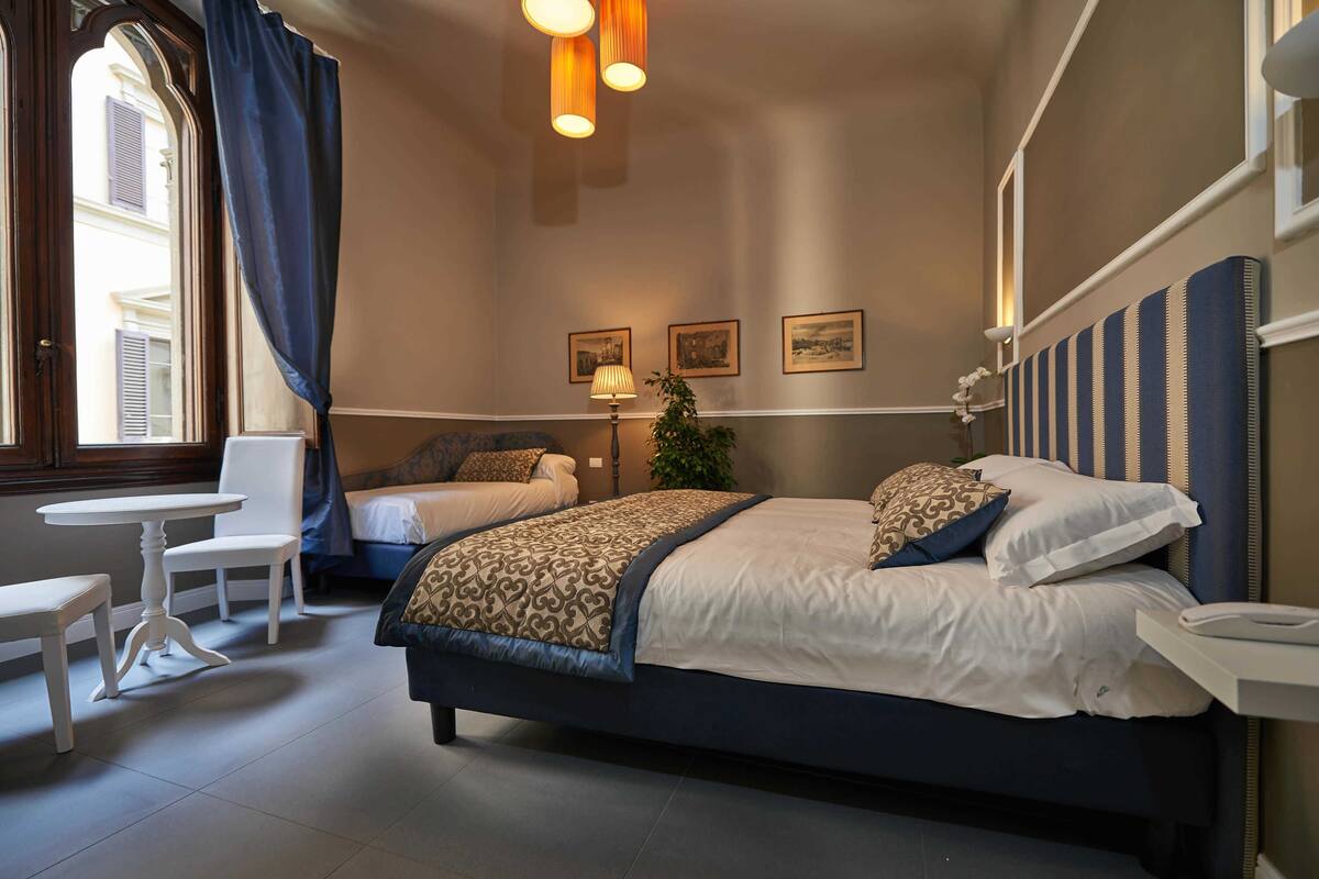 A hotel room with a large bed, a smaller bed, a desk, and chairs. There are three framed pictures on the wall and a plant on the desk.