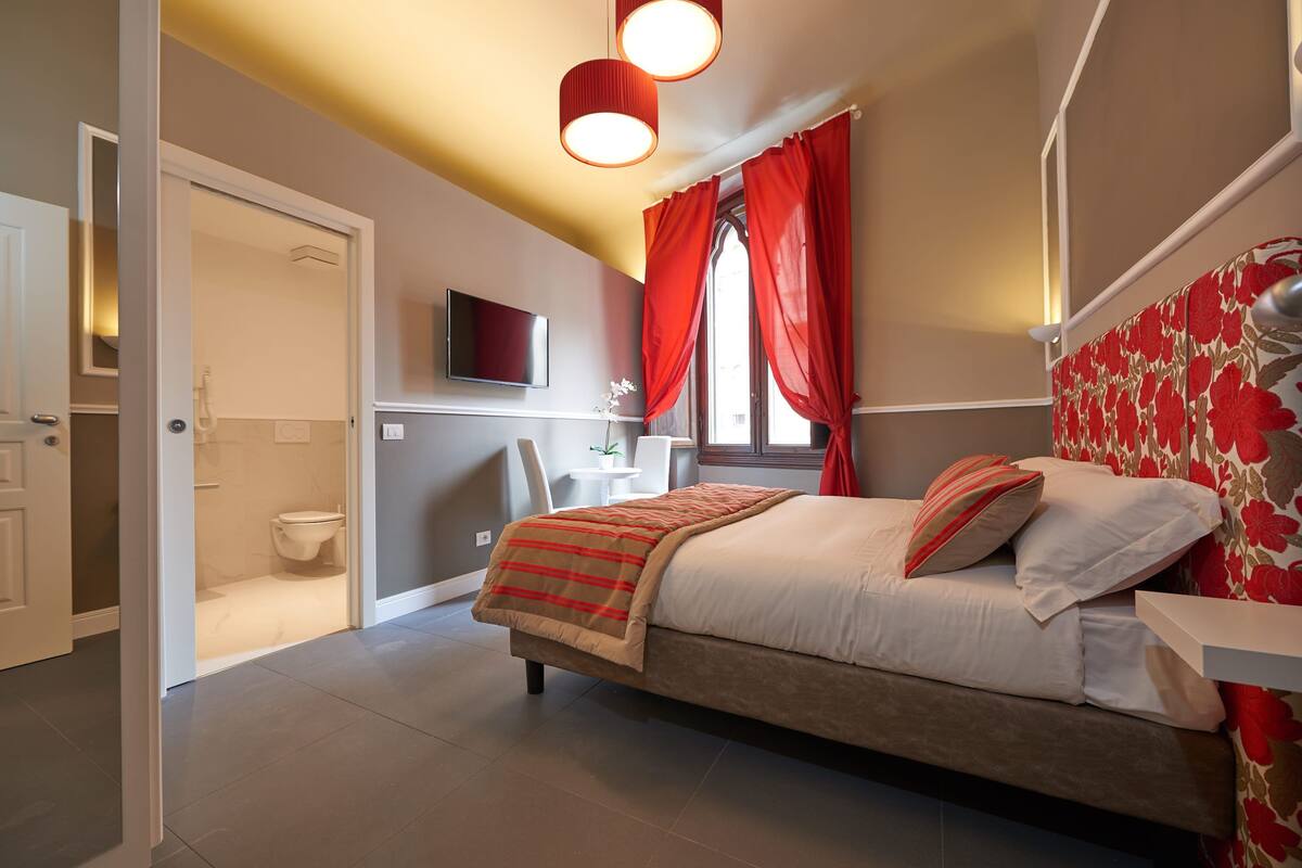 A bedroom with a bed, a TV mounted on the wall, a bathroom with a toilet, and red curtains.