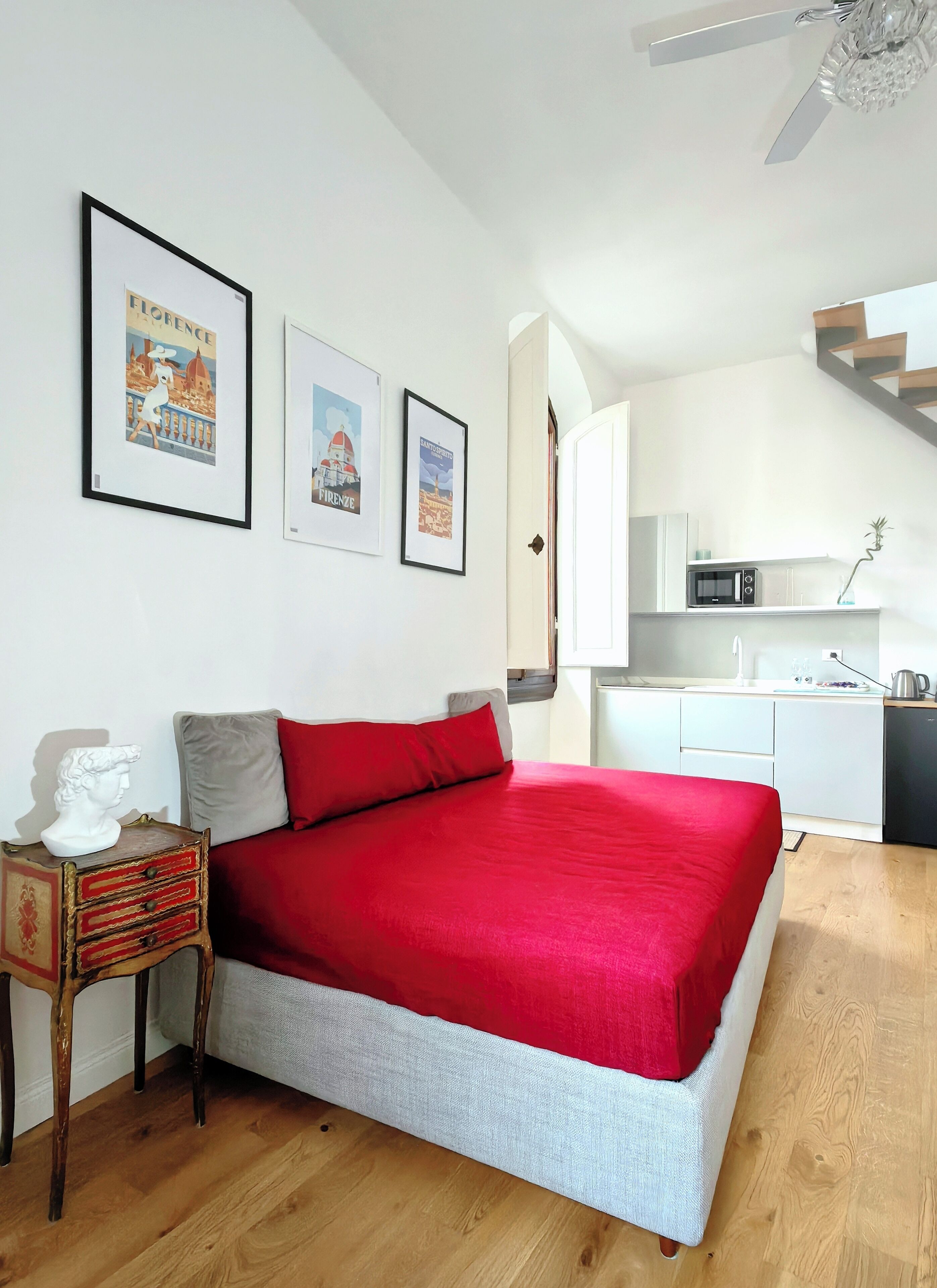 A bedroom with a red bedspread, a small bedside table with a bust sculpture, and framed travel posters on the wall.