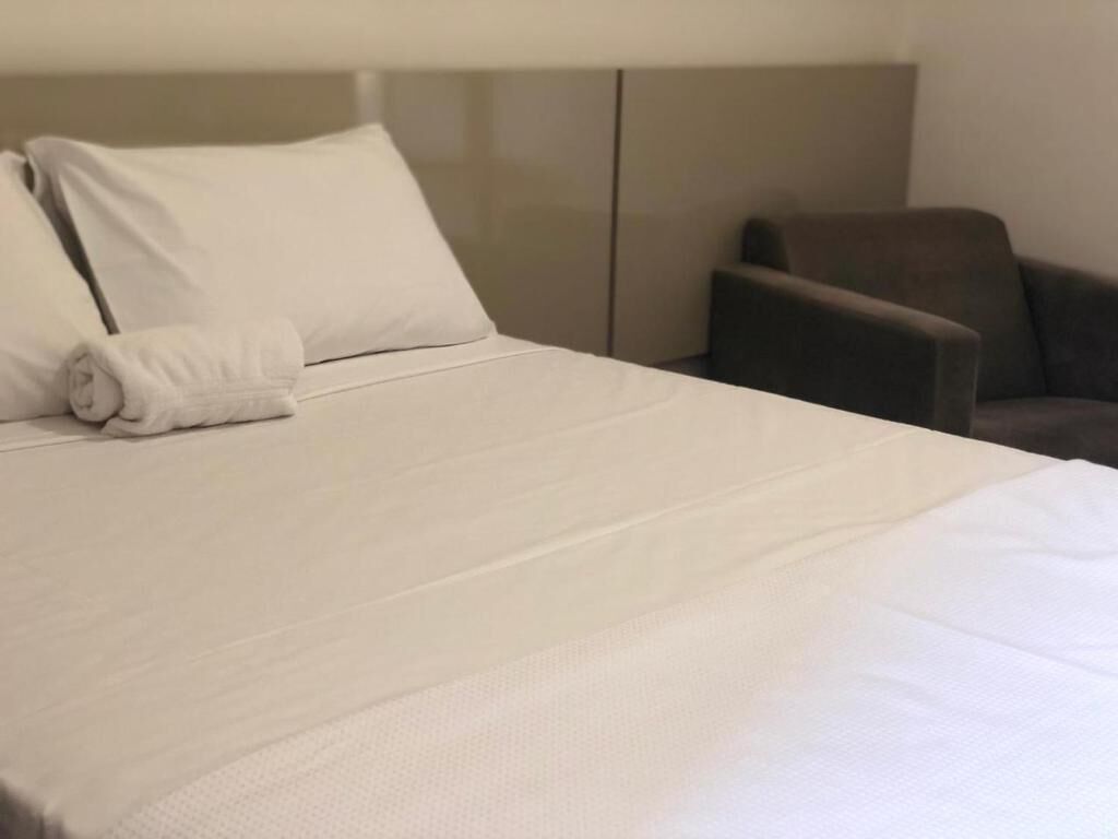 Siististi pedattu sänky valkoisilla lakanoilla ja rullalle taitettu pyyhe, tummanvärinen nojatuoli ja pelkistetty seinä hotellihuoneessa.