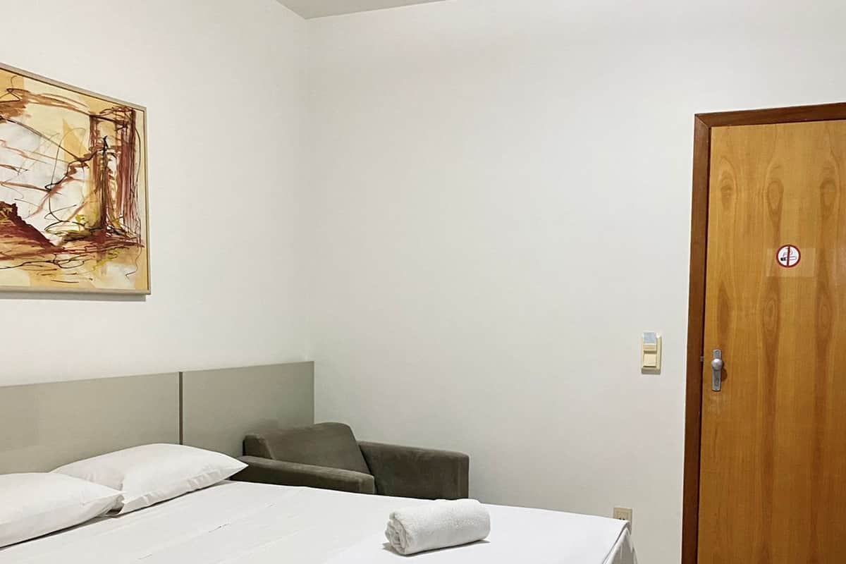 Una habitación de hotel con una cama, una silla, una puerta de madera y un cuadro en la pared.