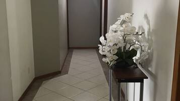Um corredor com piso de azulejos, uma mesa de madeira com arranjo de flores brancas e uma porta no final.