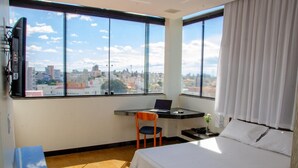 Une chambre d’hôtel équipée d’un lit, d’un bureau avec un ordinateur portable, d’une chaise, d’une télévision et d’une grande fenêtre donnant sur la ville.