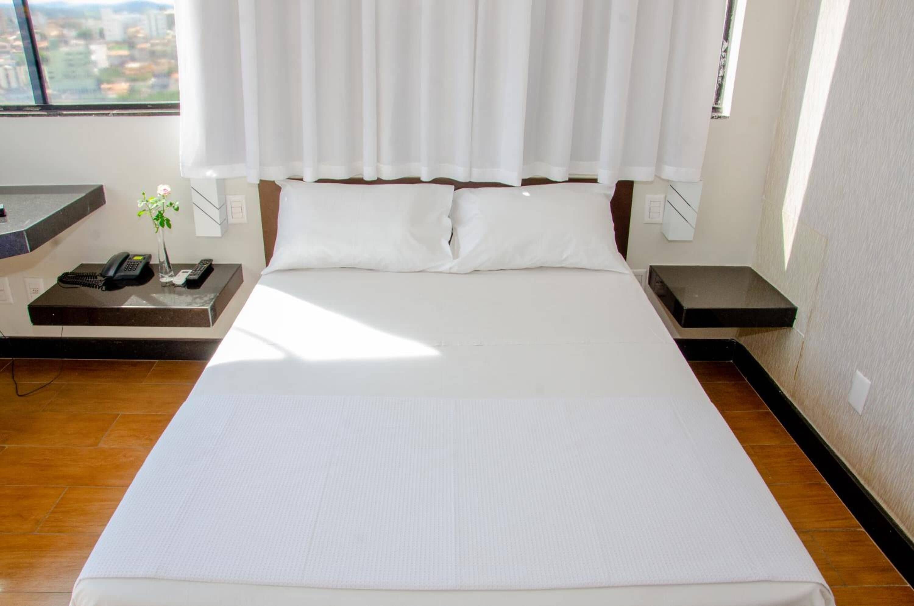 A hotel room with a large bed, a bedside table with a phone and remote controls, a small vase with a flower, and a view of the city through the window.