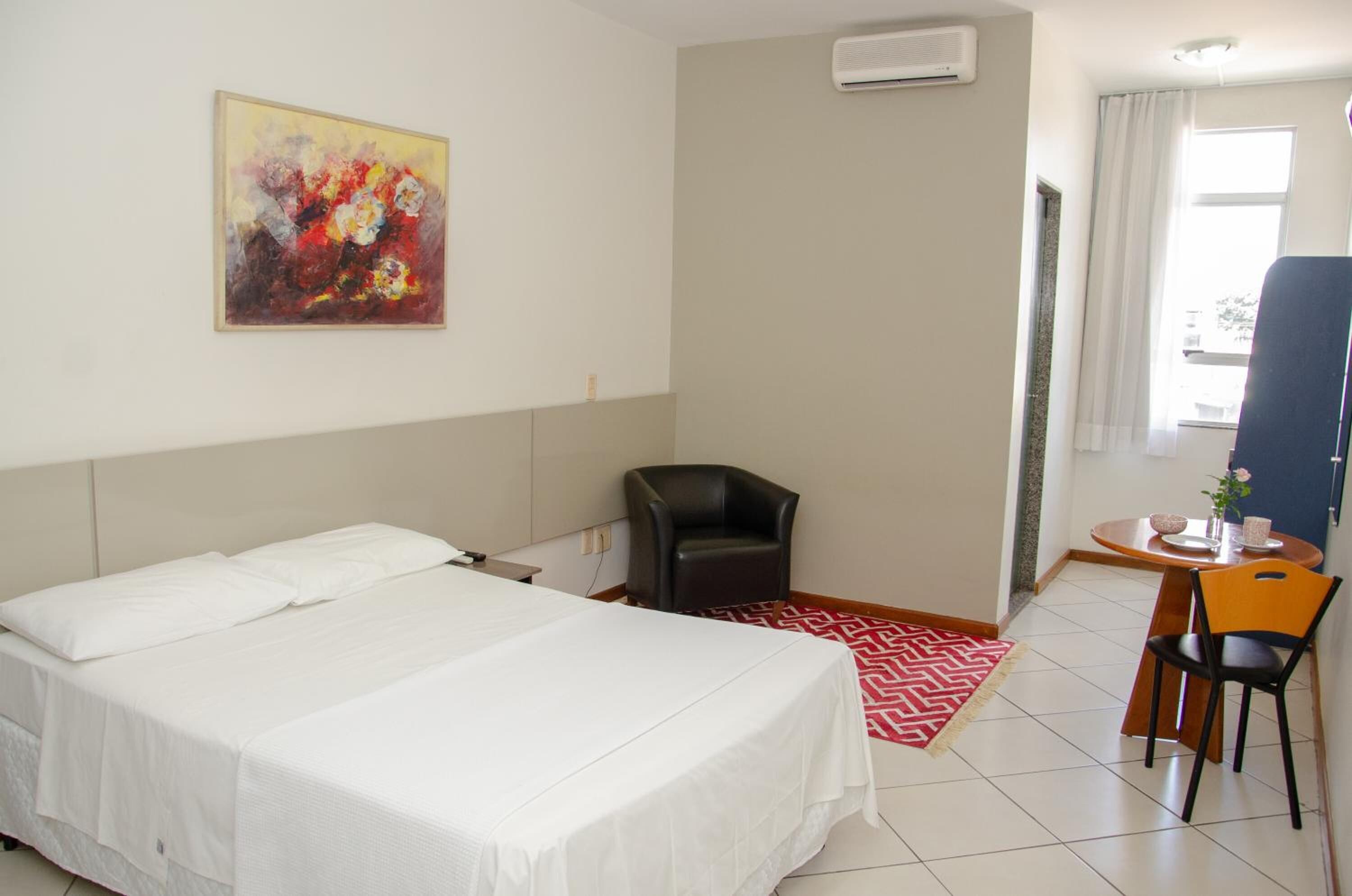 A hotel room with a bed, a desk with a chair, a small table with a vase, and a painting on the wall.