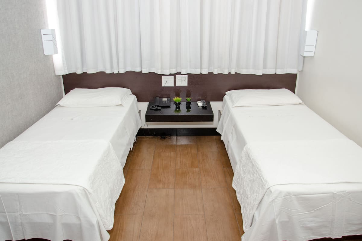Habitación de hotel con dos camas individuales, una mesita con un teléfono y una planta, y una ventana con cortinas blancas.