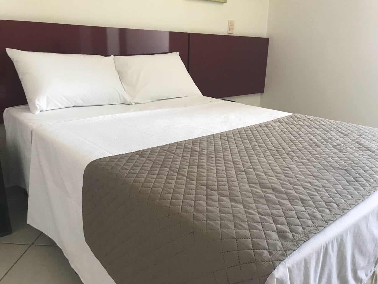 Una cama con ropa de cama blanca y una cubierta acolchada marrón, un cabecero oscuro y una pared lisa con un interruptor de luz.