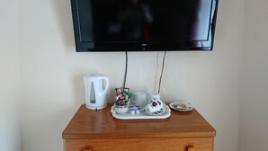 Uma cômoda de madeira com uma televisão de tela plana presa acima. Sobre a cômoda, há uma chaleira elétrica branca, um suporte com xícaras e um bule, e um prato com um biscoito.
