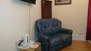 Uma sala de estar com um sofá de couro azul, uma televisão fixada na parede, um quadro emoldurado e uma mesinha com uma chaleira e chávenas.