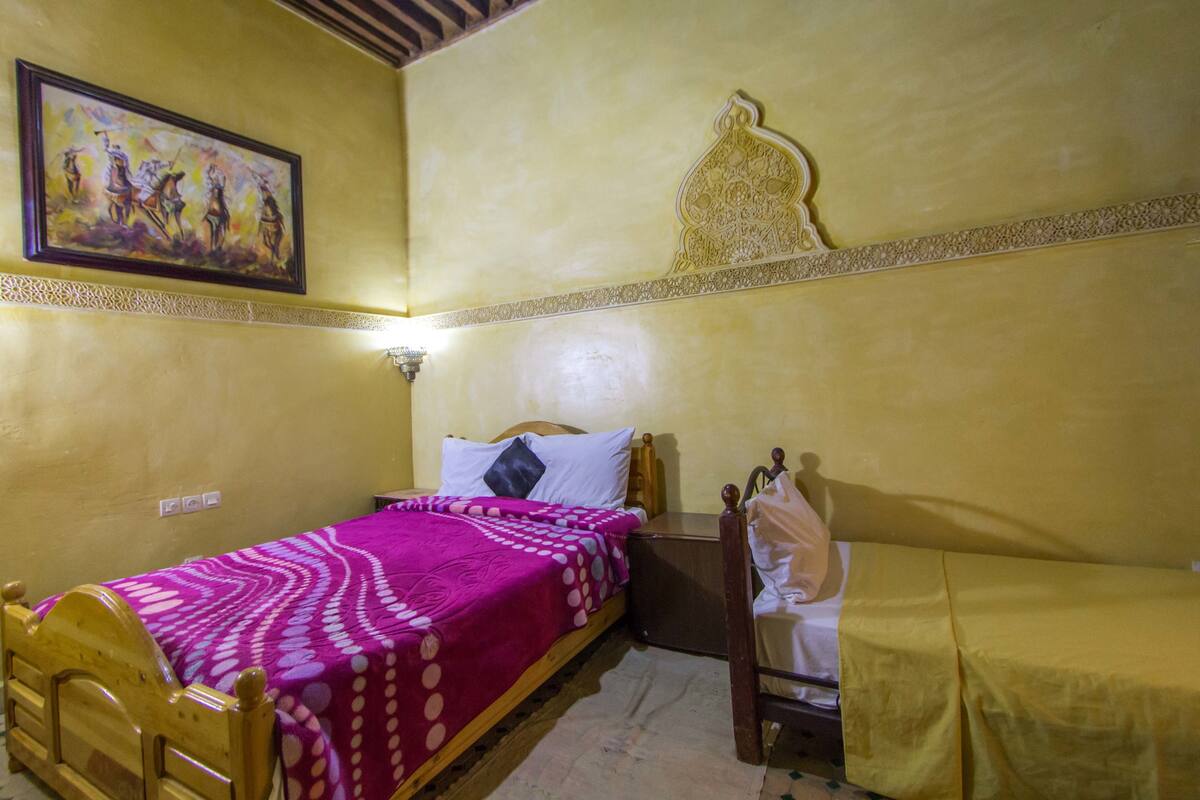 A room with a single bed, a chair, a nightstand, and a wall-mounted lamp.