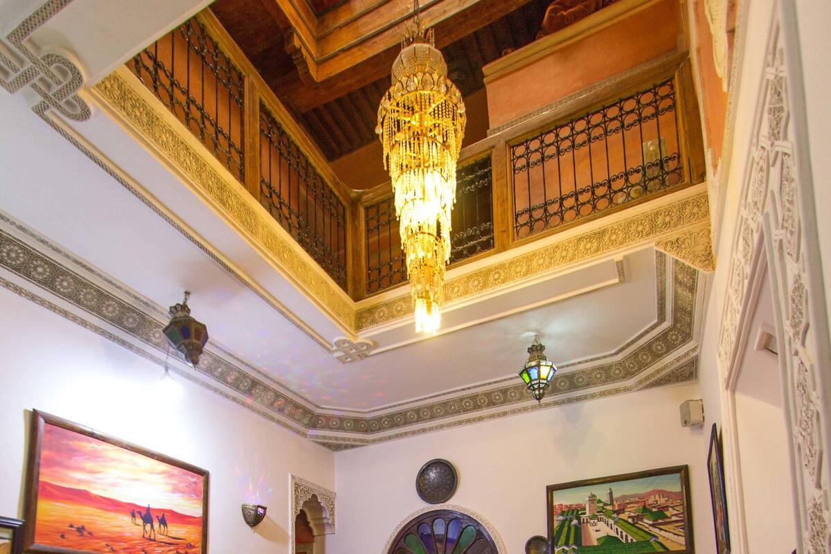 A traditional interior with a high ceiling, ornate wooden beams, a chandelier, a sofa set, a table with a bottle, and a patterned floor.