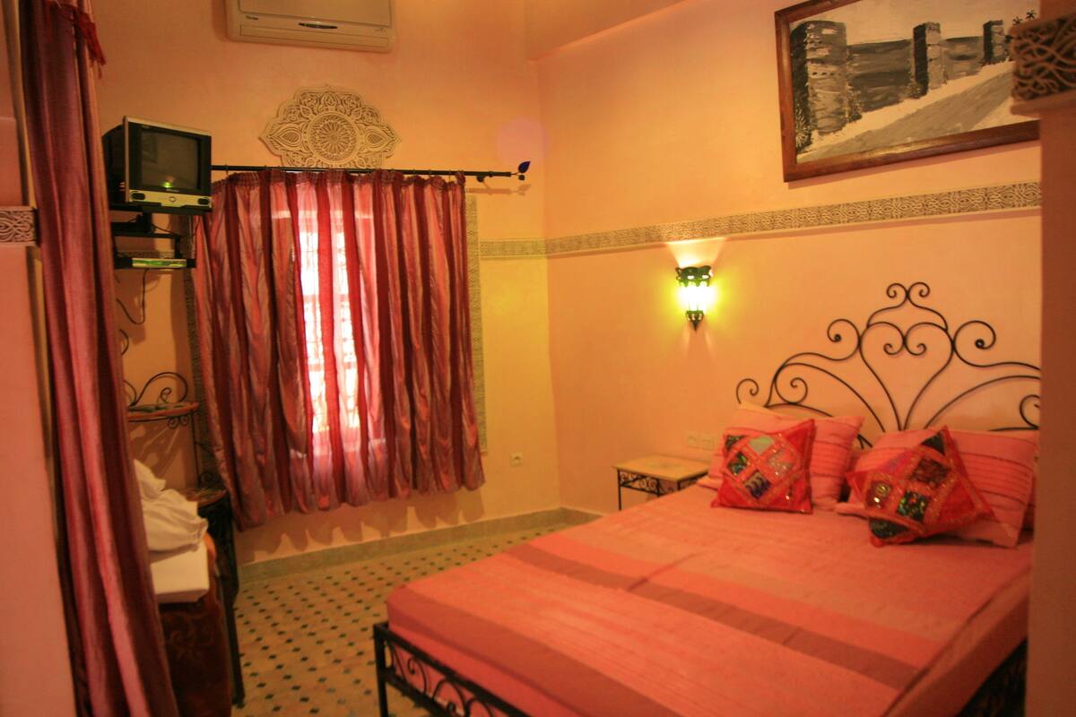 A bedroom with a bed, curtains, a wall-mounted light, a small table, and a decorative wall piece.
