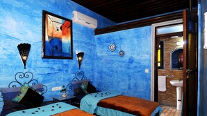 A room with two beds, a wall-mounted air conditioner, a small table with a vase, and a bathroom visible through an open door.