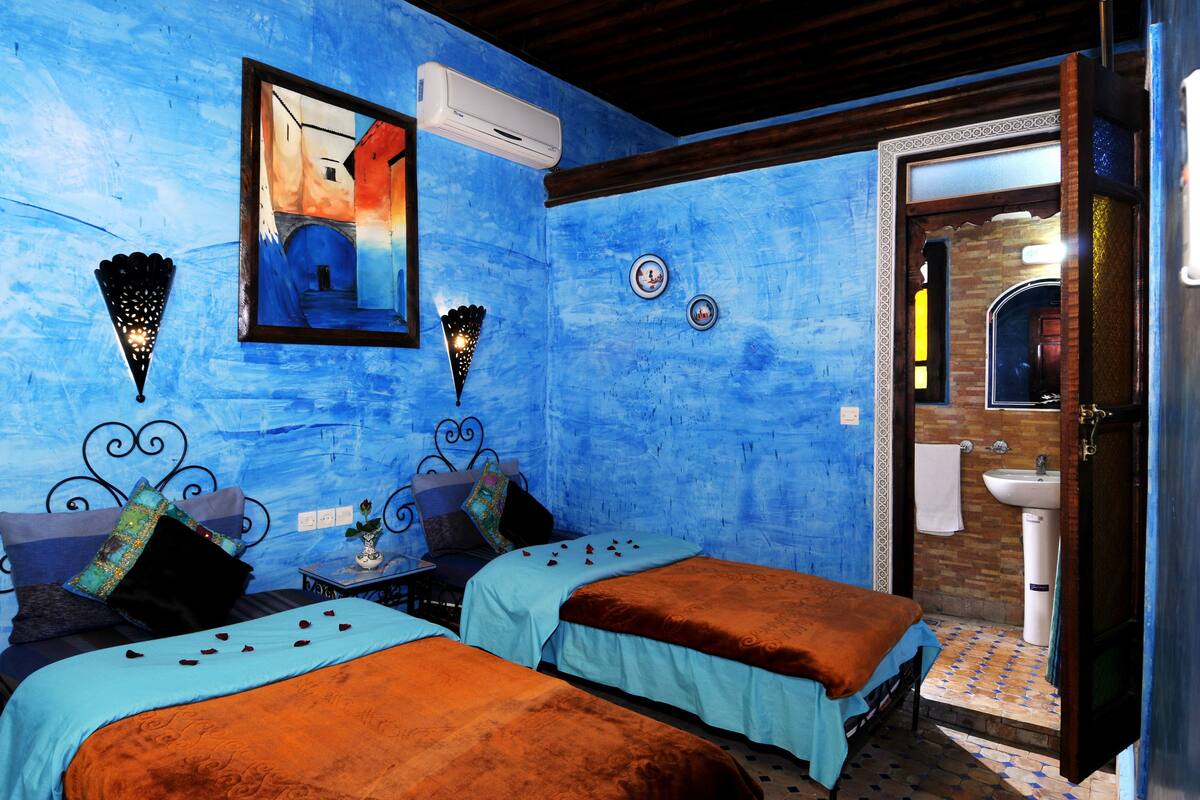A room with two beds, a wall-mounted air conditioner, a small table with a vase, and a bathroom visible through an open door.