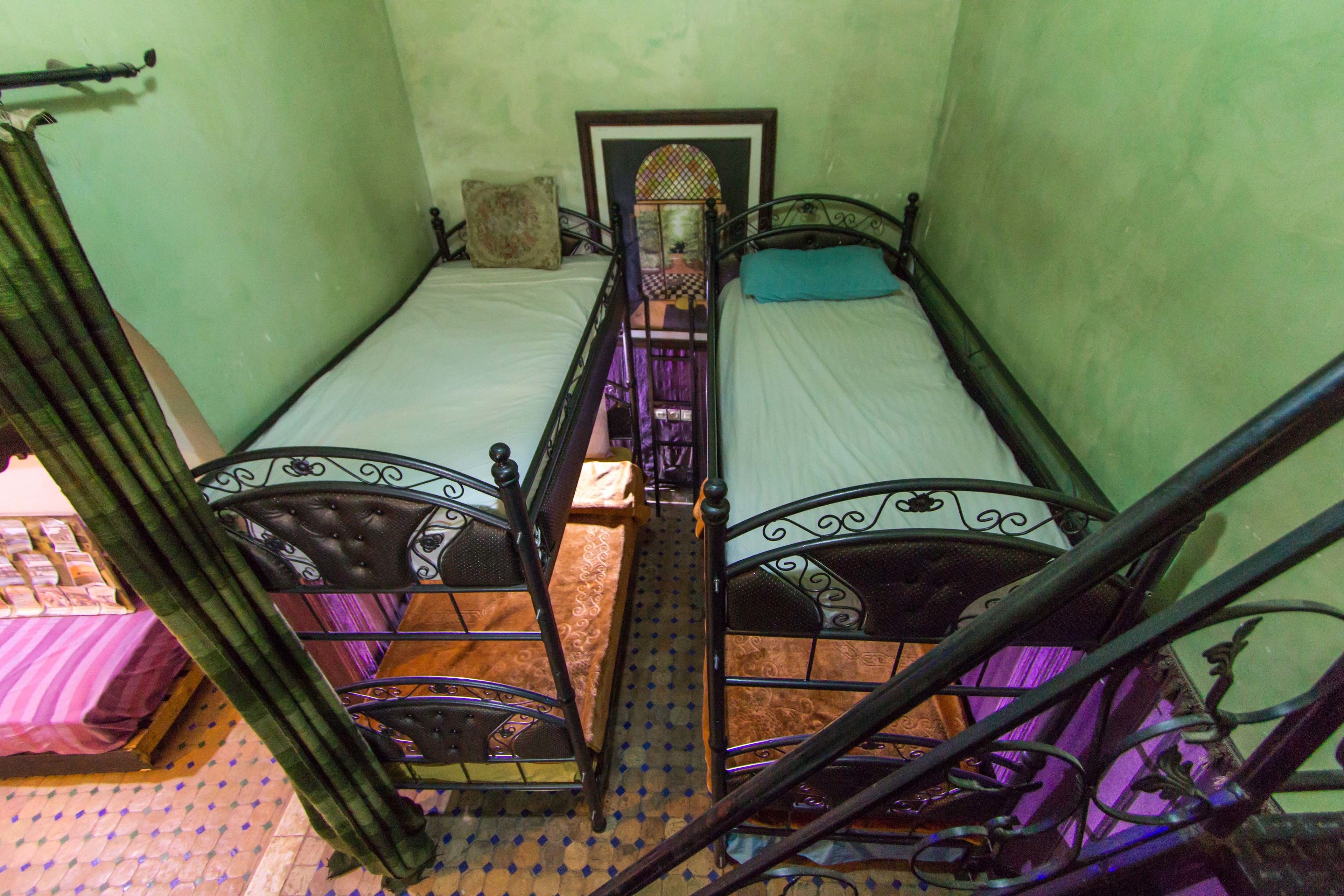 Quarto pequeno e estreito com duas camas de estrutura metálica, uma escada de madeira e piso estampado.