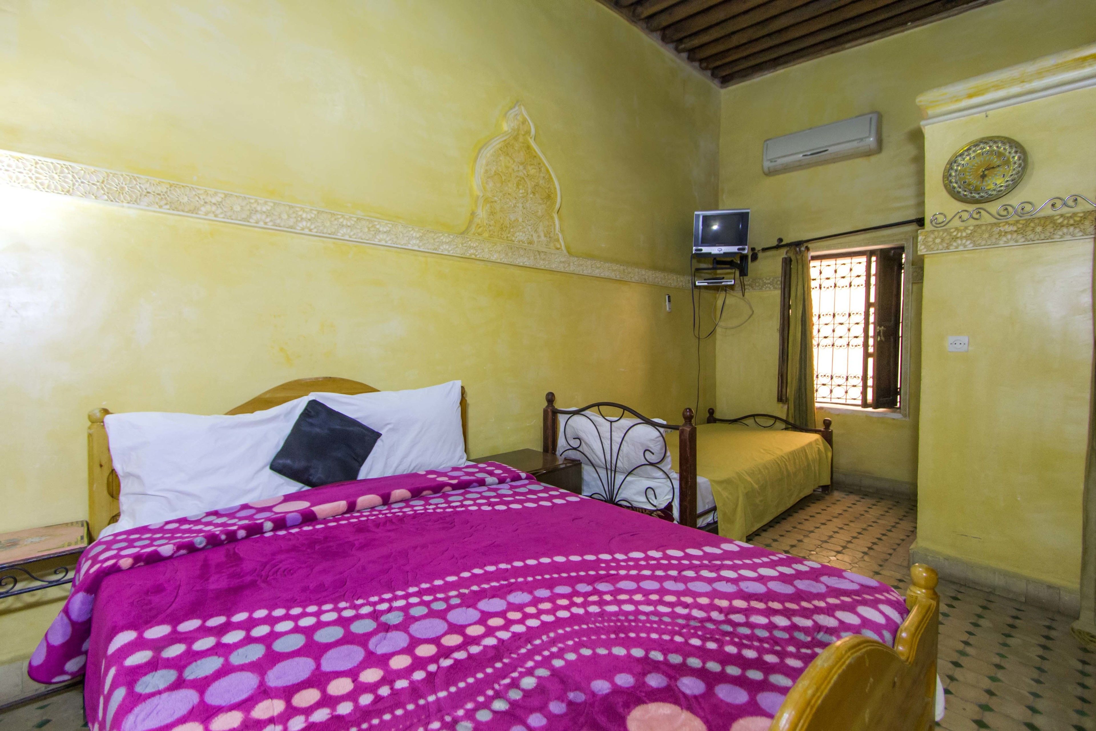 A bedroom with a bed, a bedside table, a wall-mounted air conditioner, a window with bars, and a decorative clock.