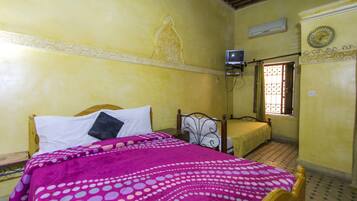 A bedroom with a bed, a bedside table, a wall-mounted air conditioner, a window with bars, and a decorative clock.