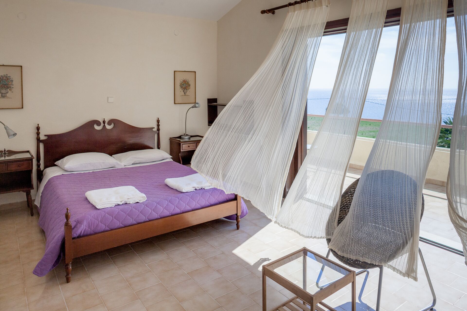 A bedroom with a bed, a nightstand, a lamp, a chair, and a view of the sea through sheer curtains.
