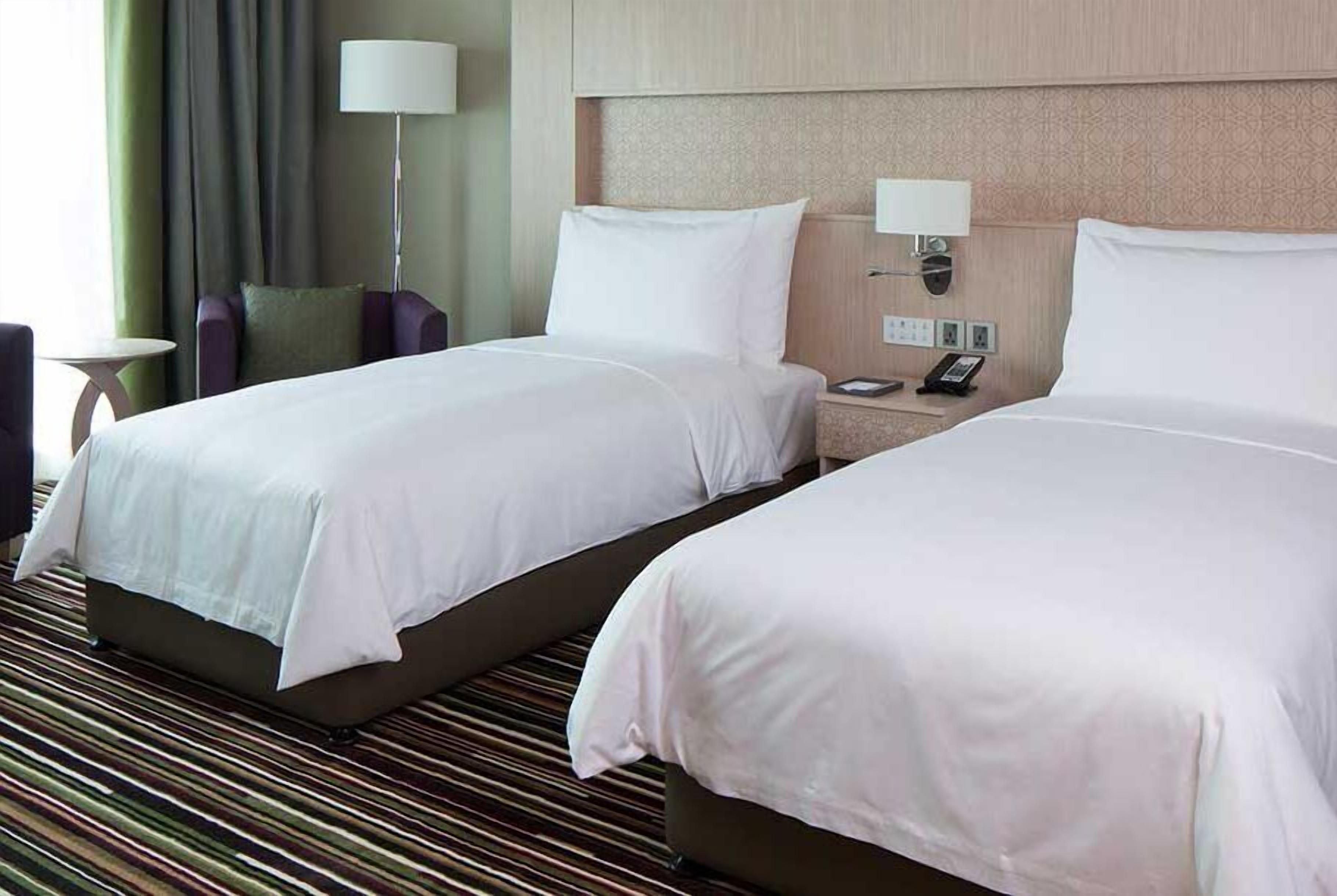 A hotel room with two beds, a bedside table with a lamp, and a wall-mounted phone.